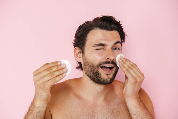 White shirtless man winking while doing makeup with cotton pads isolated over pink background - Zdjęcie, obraz