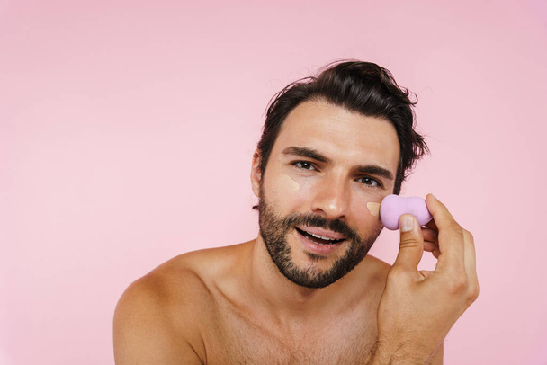 White shirtless man applying concealer with sponge isolated over pink background - Foto, Imagem