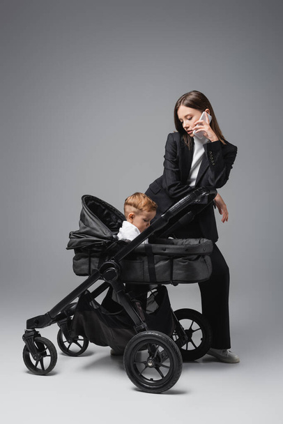 businesswoman talking on smartphone near toddler son in pram on grey - Photo, image