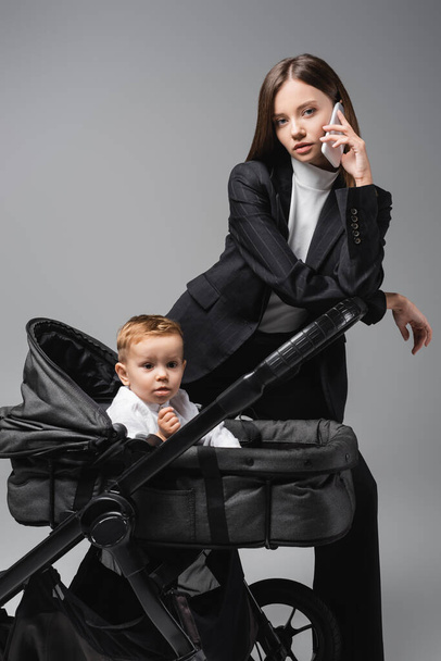 woman in suit talking on mobile phone near toddler son in baby carriage isolated on grey - Fotografie, Obrázek