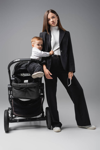 businesswoman holding smartphone with blank screen near pram with son on grey - Photo, Image