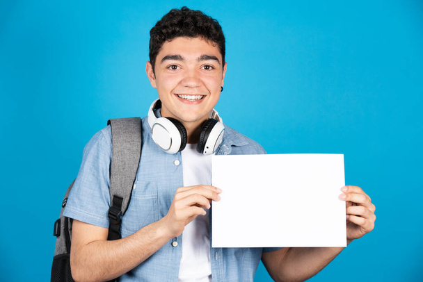 Hispanic college student holding white sheet of paper with copy space and looking at camera isolated on blue background. - Photo, image
