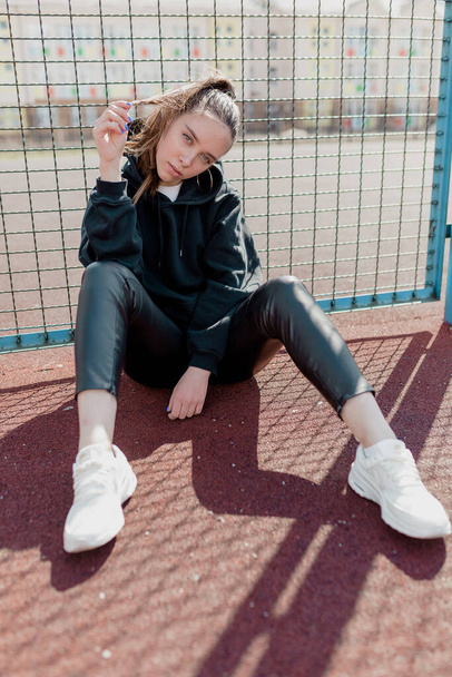 Full-length φωτογραφία του αρκετά ευρωπαϊκό κορίτσι σε μαύρο casual ρούχα κοιτάζει κάμερα και αγγίζοντας τα μαλλιά της στην πλατεία αθλητισμού. Υψηλής ποιότητας φωτογραφία - Φωτογραφία, εικόνα