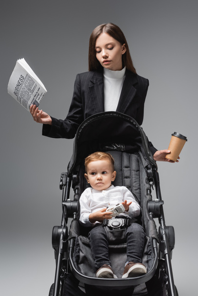 businesswoman with newspaper and coffee to go near son sitting in pram with toy car isolated on grey - Fotografie, Obrázek