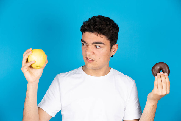 Hispanic young man looking with repugnance at apple while holding a chocolate donut or doughnut in the other hand isolated on blue background - Photo, Image