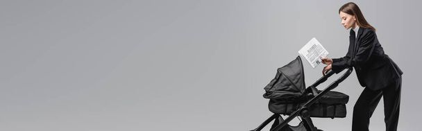 boční pohled na ženu v černém obleku držící obchodní noviny poblíž kočárku izolovaného na šedé, banner - Fotografie, Obrázek