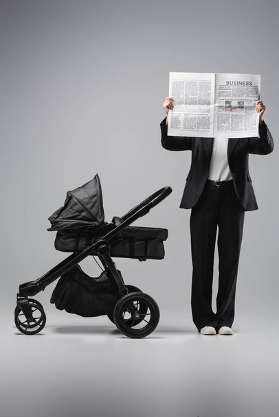 mujer en traje negro oscureciendo la cara con periódico cerca de carro de bebé en gris - Foto, imagen