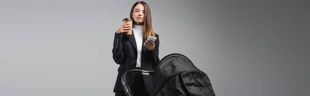 donna con smartphone e caffè per andare a guardare la macchina fotografica vicino alla carrozzina isolata su grigio, banner - Foto, immagini