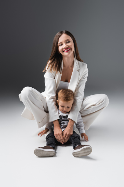 cheerful woman looking at camera and embracing son sitting on grey - Fotoğraf, Görsel