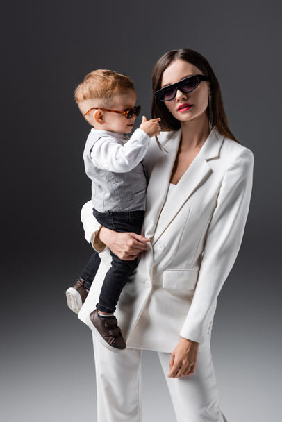 trendy woman in sunglasses holding little son and looking at camera on grey - Photo, Image