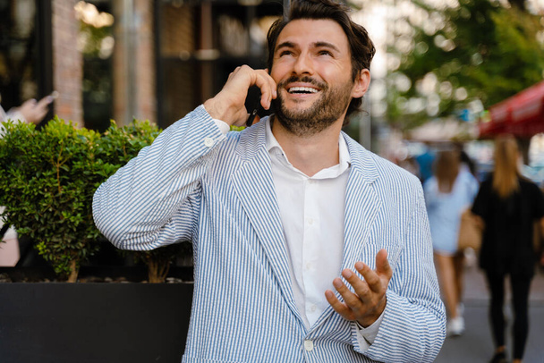 Молодой человек жестикулирует и разговаривает по мобильному телефону, стоя на улице - Фото, изображение