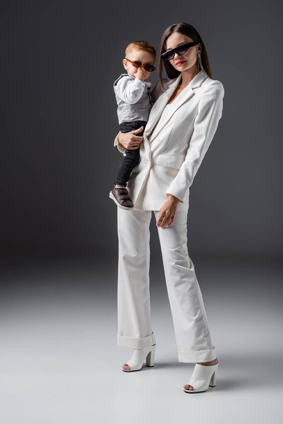 full length view of woman in trendy suit holding little son adjusting sunglasses on grey - Φωτογραφία, εικόνα