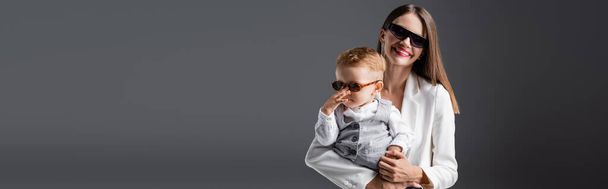 pleased woman in white blazer holding son touching trendy sunglasses isolated on grey, banner - Foto, Imagen