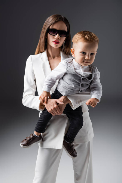 woman in sunglasses holding son wearing stylish clothes on grey - Foto, imagen