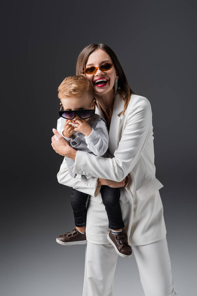 laughing woman holding son adjusting stylish eyeglasses on grey - Foto, Bild
