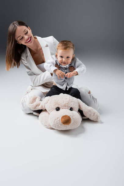 young woman in white suit playing with little kid sitting on toy dog on grey - Fotoğraf, Görsel