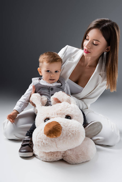 baby boy looking at camera while sitting on toy dog near mother on grey - Photo, Image