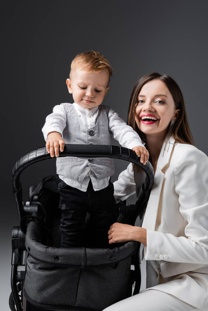 mujer alegre mirando la cámara cerca de hijo con estilo en carro de bebé en gris - Foto, imagen