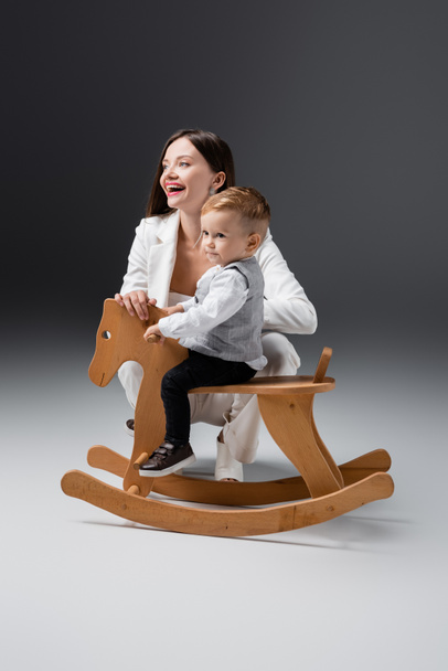 cheerful young woman smiling near son riding rocking horse on grey - Foto, Imagen