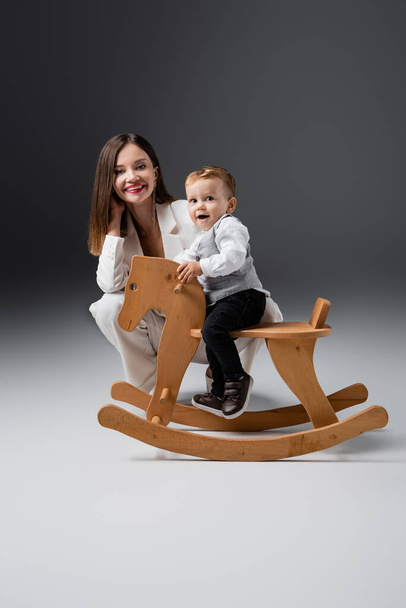 amazed boy riding rocking horse near happy young mother on grey - Foto, immagini