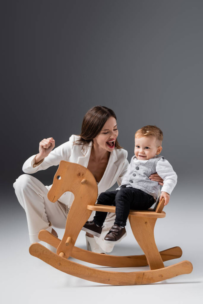 excited woman showing win gesture near son riding rocking horse on grey - Photo, Image