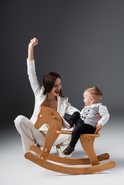excited mother showing triumph gesture near little son on rocking horse on grey - Фото, изображение