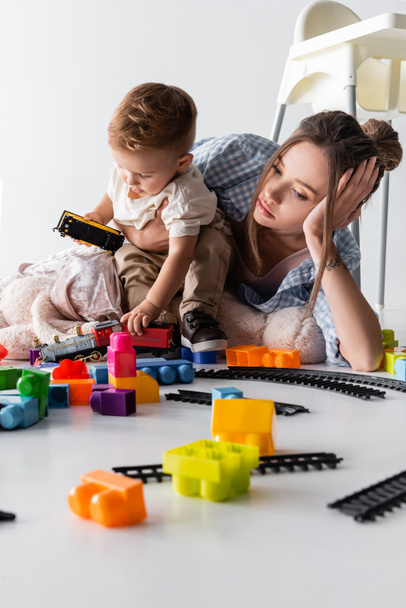 tired mother lying near toddler son playing with toy train on white - Photo, Image