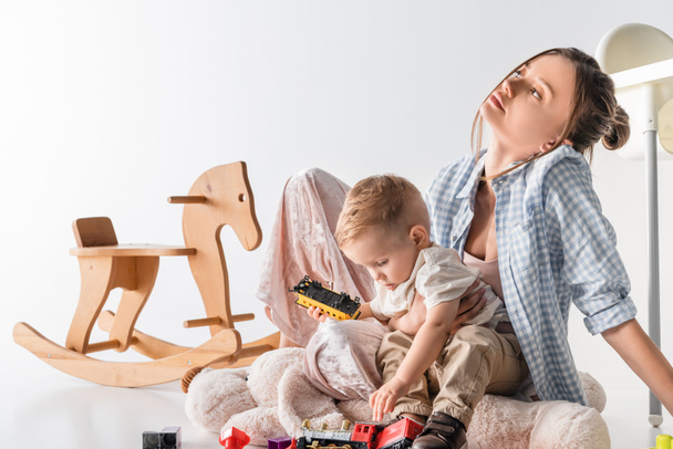 tired woman sitting near playing son and rocking horse on white - Foto, Imagem