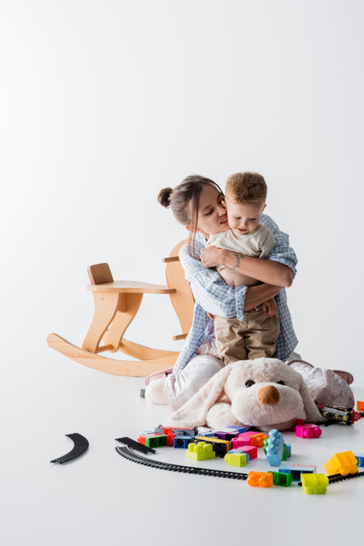 mujer emocionada abrazando hijo cerca de juguetes y balanceo caballo en blanco - Foto, imagen