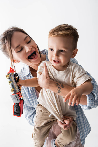 cheerful woman embracing toddler son holding toy train isolated on white - Foto, Imagem
