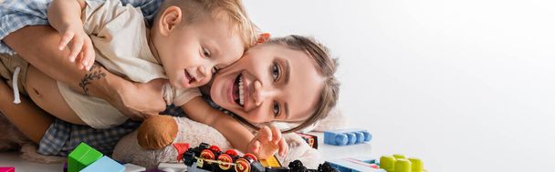 cheerful woman looking at camera while playing with son on white, banner - Foto, imagen