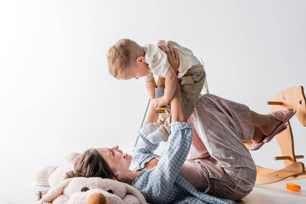 cheerful woman lying on toy dog and playing with toddler son on white - Foto, Imagem