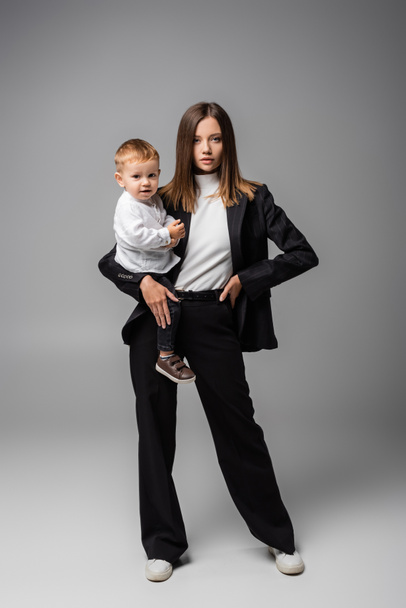 businesswoman in black suit standing with toddler son on grey - Φωτογραφία, εικόνα