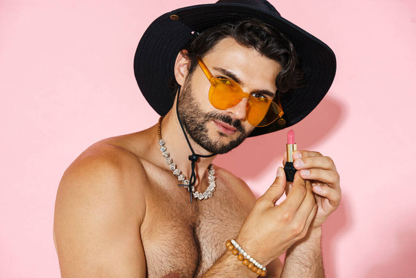 Young shirtless man showing lipstick and looking at camera isolated over pink background - Foto, imagen