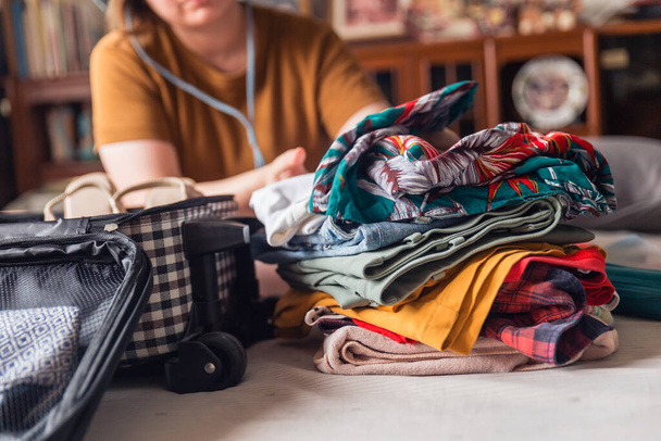 女性は選択された所持品の山から服を折り、ベッドの上に荷物に入れます。休暇に行くためにパッキング。服を中心に. - 写真・画像