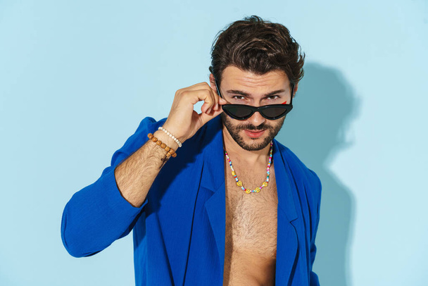 Young man wearing sunglasses posing and looking at camera isolated over blue background - Fotografie, Obrázek