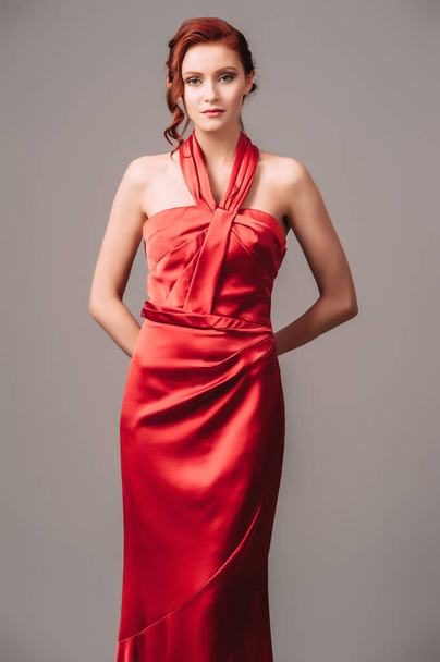 Bright young lady in red sleeveless dress with gathers and a high halter neckline on grey studio background. Natural makeup and hairdo concept. Candid minimalistic portrait. - Foto, Imagem
