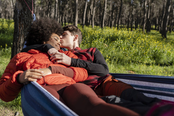 Χαριτωμένο πολυφυλετικό ζευγάρι φιλιά σε άνετο αιώρα σε εξωτερικούς χώρους κοντά στο δάσος, όμορφος άντρας αγκαλιάζει γυναίκα, ενώ χαλαρώνει στις διακοπές - Φωτογραφία, εικόνα