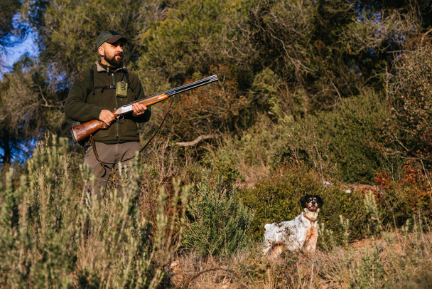 Хантер держит пистолет посреди природы и смотрит в сторону со своей собакой. - Фото, изображение
