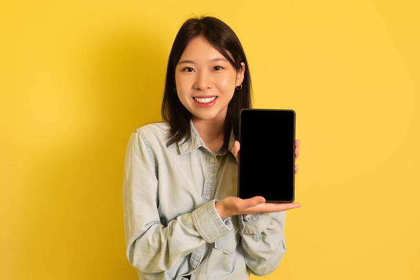 Millennial Asian lady presenting mockup for advertisement on tablet screen over yellow studio background - Photo, Image
