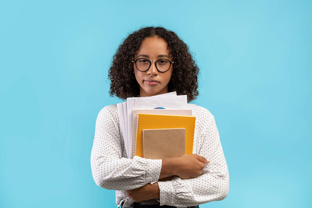 Dubbia giovane studentessa nera con quaderni e carte guardando la fotocamera su sfondo blu studio - Foto, immagini