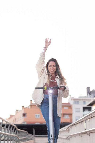 Vertical shot of young woman having fun with her electric scooter around the city. Arm raised as a happiness sign - Foto, Imagen