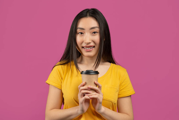 Szczęśliwa koreańska pani trzyma kawę na wynos w jednorazowym papierowym kubku i uśmiecha się do kamery, pozując na fioletowym tle - Zdjęcie, obraz