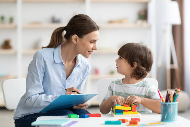 Professional woman psychologist exercising with little boy, checking readiness for school and taking notes at office - Photo, Image