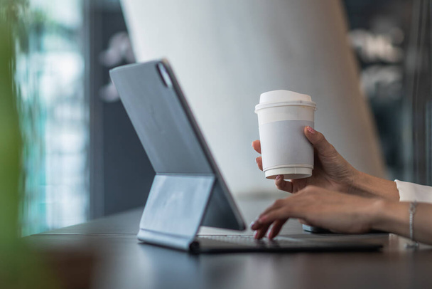 Una mujer de negocios sostiene una taza de papel de café caliente y está utilizando una tableta conectada a Internet para intercambiar acciones a través de la aplicación durante un descanso en una cafetería. - Foto, Imagen