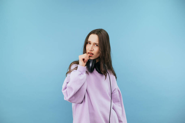 Nervous brunette girl in casual clothes and headphones bites her nails and looks at the camera with a sad face. - Fotoğraf, Görsel