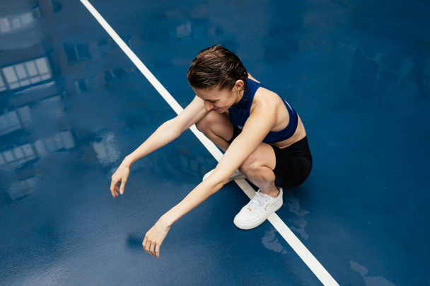 Pozitif spor kadını spor sahasında sokakta antrenman yapıyor, mavi ıslak bir yüzeye oturuyor ve gülümsüyor.. - Fotoğraf, Görsel