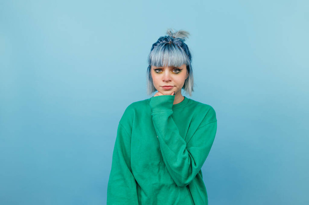 Pensive woman in green sweatshirt and with blue hair isolated on blue background with serious face. - Zdjęcie, obraz