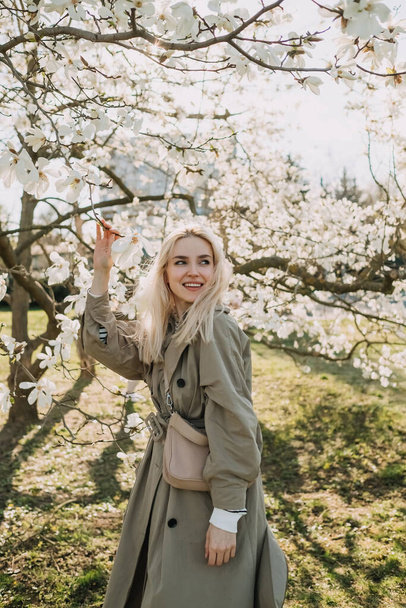 Young happy blonde woman posing under a magnolia tree in bloom, a sunny spring day, in a park. - Foto, Imagen