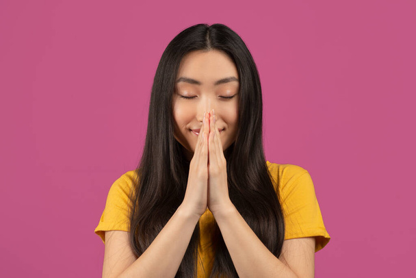 Mírumilovná asijská dáma modlící se se zavřenýma očima, držící se za ruce v prosebném gestu, stojící na fialovém pozadí - Fotografie, Obrázek
